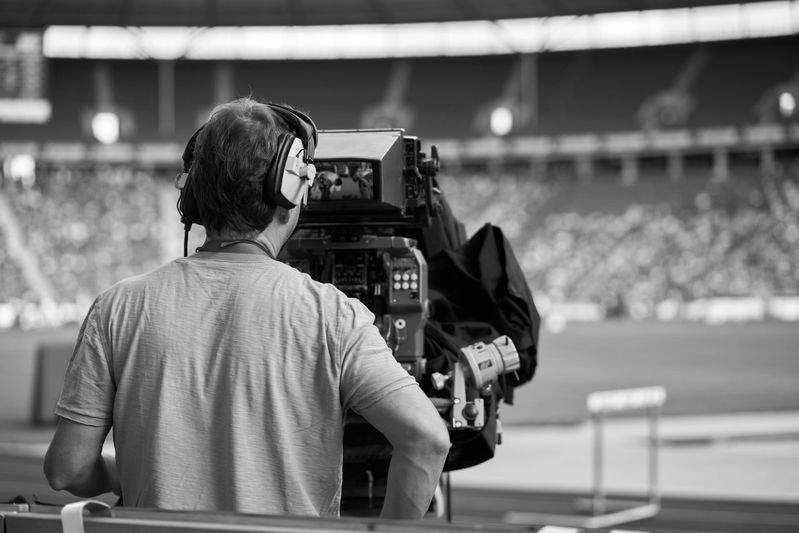 Cameraman wearing headphones operates a large video camera in a stadium, capturing an event with blurred spectators in the background.