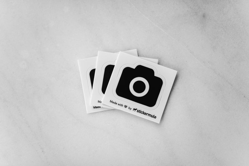 Three square stickers with a black camera icon on a marble surface. Text below reads "Made with ❤️ by Sticker Mule."