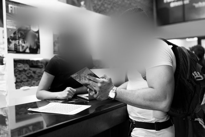 Dwie osoby przy ladzie, jedna trzyma broszurę, druga papierek. Obie twarze są rozmyte. Zdjęcie czarno-białe.