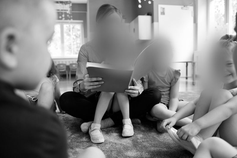 Un groupe d’enfants est assis au sol autour d’un adulte lisant un livre dans une salle de classe. L’image est en noir et blanc.