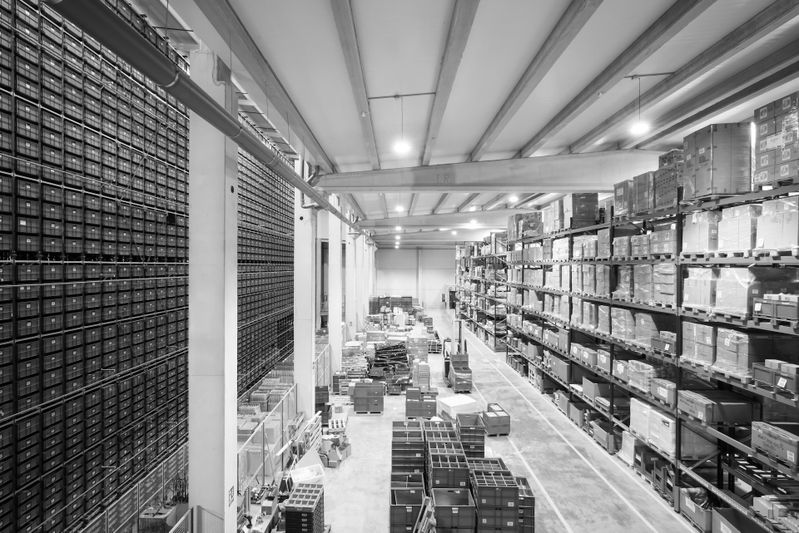 Black-and-white wide view of a large warehouse with tall shelves stacked with boxes and pallets lining a central aisle.