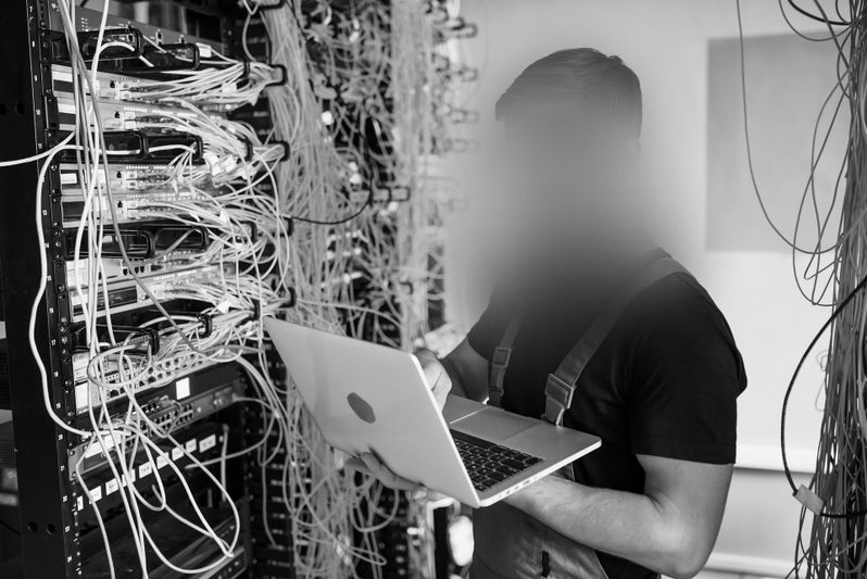 Person, die mit einem Laptop in einem Serverraum arbeitet, umgeben von Racks und verhedderten Kabeln.