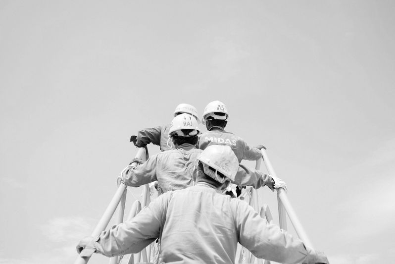 Fotografía en blanco y negro de cuatro trabajadores con cascos blancos y monos subiendo unas escaleras.