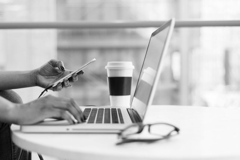 Persona que usa una computadora portátil y un teléfono inteligente en una mesa redonda con una taza de café para llevar y vasos cerca, en un entorno interior luminoso.