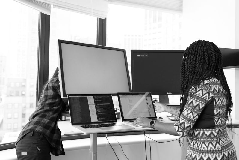 Deux personnes travaillant à des bureaux debout avec des ordinateurs portables et des écrans dans un bureau lumineux avec de grandes fenêtres.