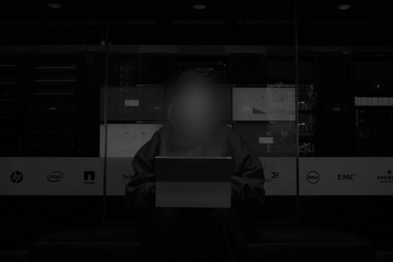 Person with blurred face using a laptop in a dimly lit server room, surrounded by various equipment and logos.