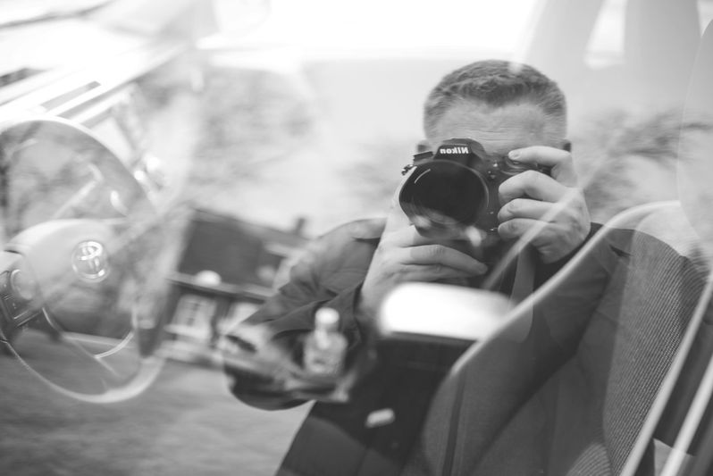 En el cristal del coche se refleja un hombre tomando una foto con una cámara Nikon, foto en blanco y negro