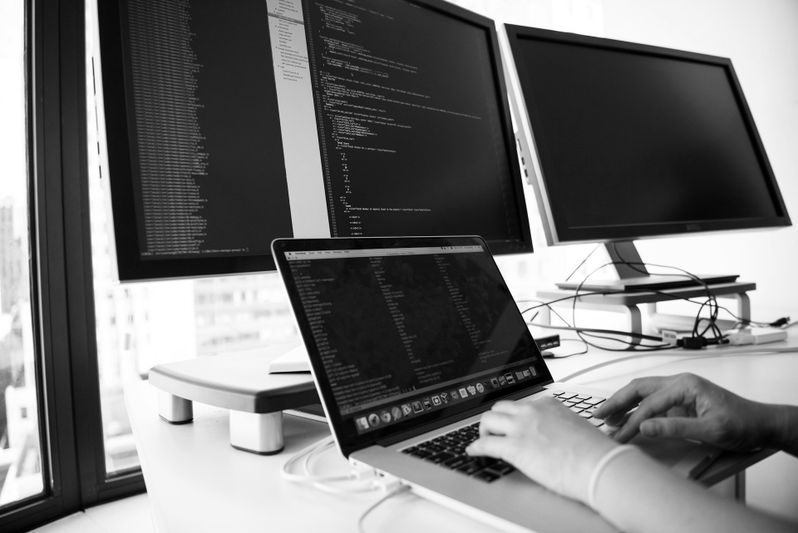 Persona escribiendo en una computadora portátil con código en la pantalla, frente a dos monitores grandes que muestran más código, en un entorno de oficina luminoso.