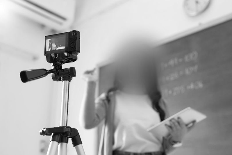 Une personne debout devant un tableau noir est filmée par une caméra sur trépied. Son visage est flou.