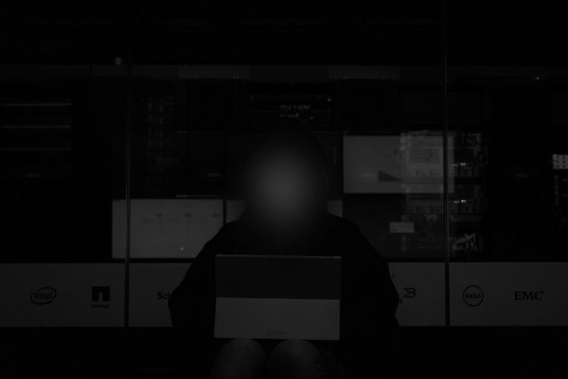 Silhouette d'une personne utilisant un ordinateur portable dans une pièce faiblement éclairée avec des serveurs informatiques et des logos en arrière-plan.