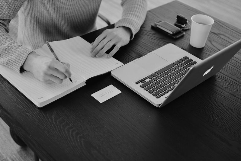 Personne écrivant dans un carnet à côté d'un ordinateur portable sur une table en bois foncé, avec une tasse et un smartphone à proximité. Image en noir et blanc.