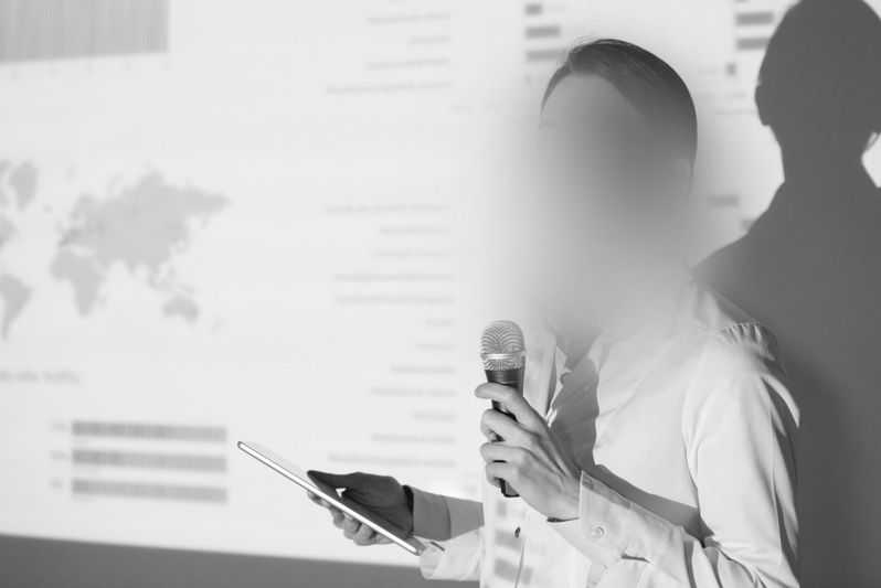 En la foto en blanco y negro se ve a una mujer con el rostro difuminado, sosteniendo una tablet y un micrófono, dando una presentación