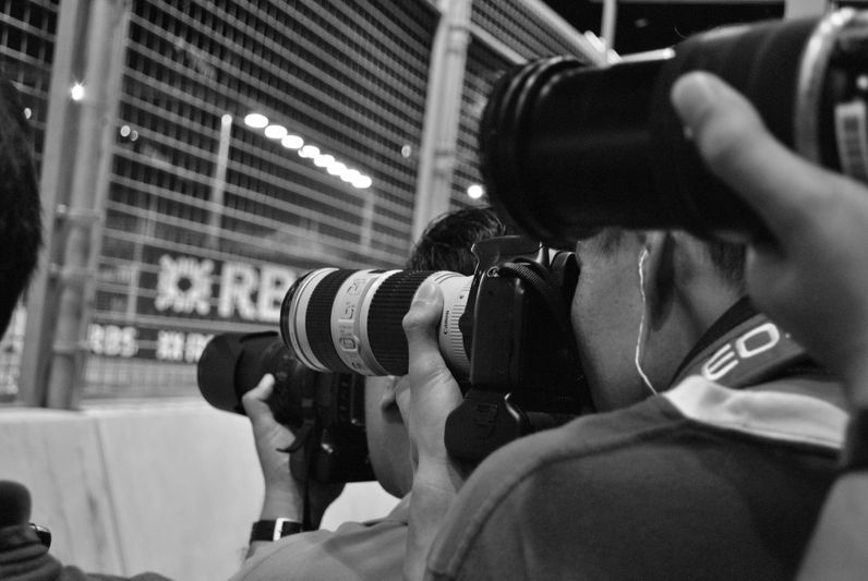Photographers with large cameras capture images at a nighttime event, standing near a wire fence.