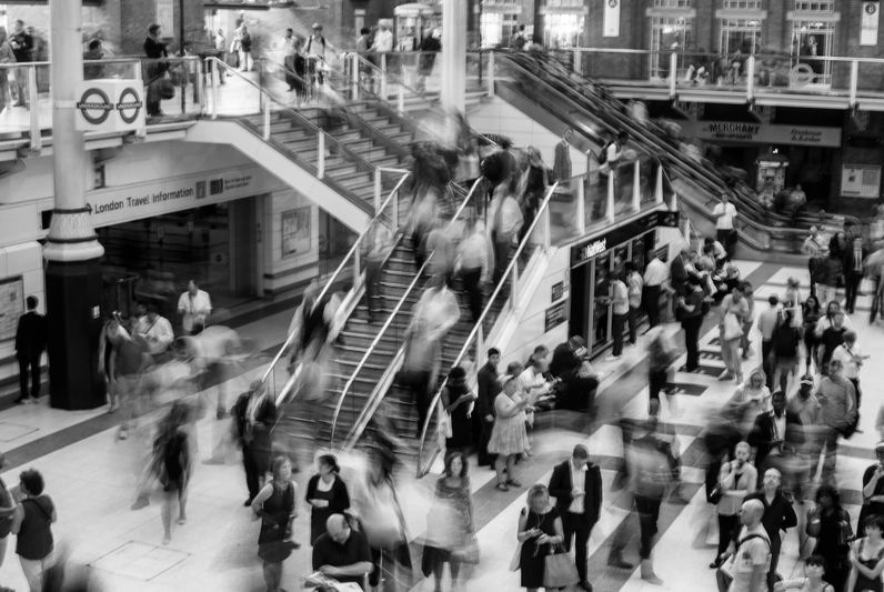 Black-and-white high-angle view of a busy train station with blurred commuters on escalators and crowds moving across the concourse.