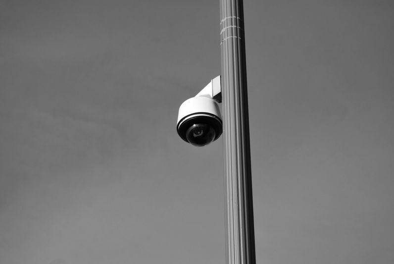 Image en noir et blanc d'une caméra de sécurité montée sur un grand poteau contre un ciel clair.