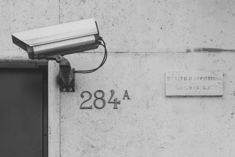 Caméra de surveillance montée au-dessus de « 284A » sur un mur portant un panneau interdisant l’affichage, en niveaux de gris.