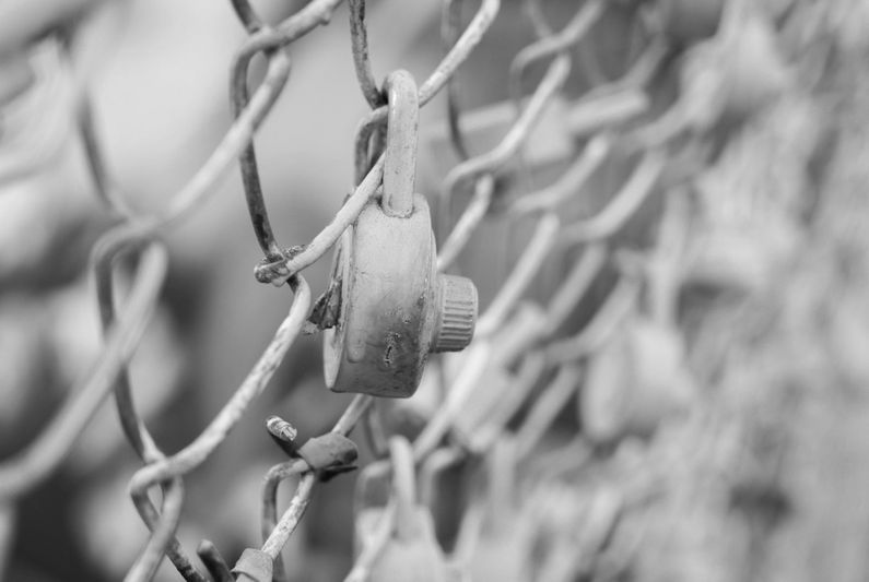 Close-up of a rusty padlock on a chain-link fence in black and white, symbolizing security and decay.