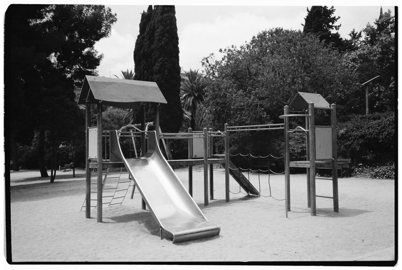 A black-and-white photo of an empty playground with a slide, climbing structures, and trees in the background.
