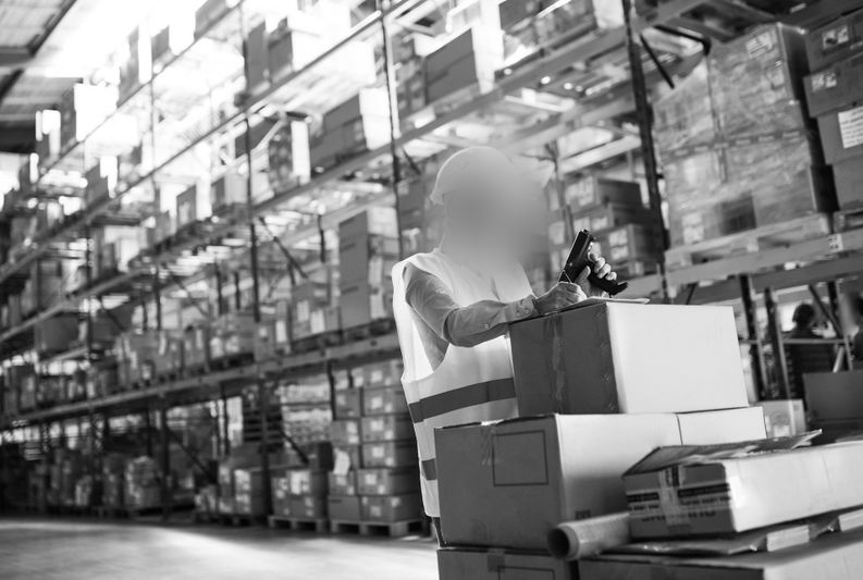 Employé dans un entrepôt scannant des cartons à l’aide d’un terminal portable, entouré de hautes étagères remplies de colis et de conteneurs.