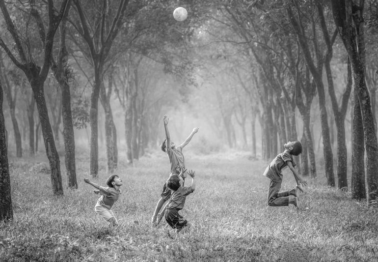 Four children joyfully jumping to catch a ball in a misty, tree-lined path. Black and white photograph capturing a moment of play.