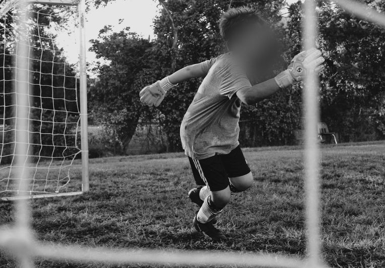 Young soccer goalie mid-action in front of a goal, wearing gloves and sportswear, with trees in the background. Black and white image.