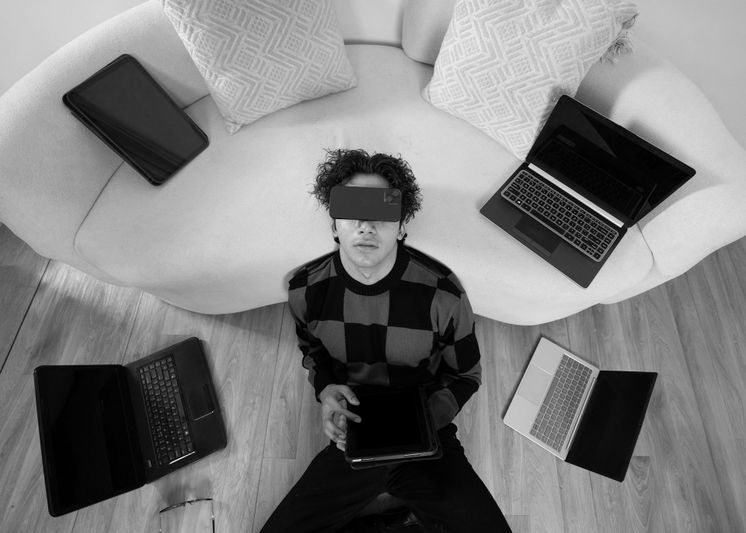Person sitting on the floor surrounded by laptops, wearing a checkered sweater, holding a tablet, and leaning against a sofa. Black and white image.