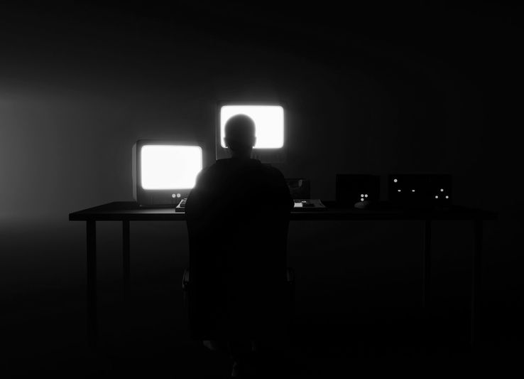 Une personne en silhouette est assise à un bureau avec des écrans d'ordinateur lumineux dans une pièce sombre, créant une atmosphère mystérieuse et axée sur la technologie.