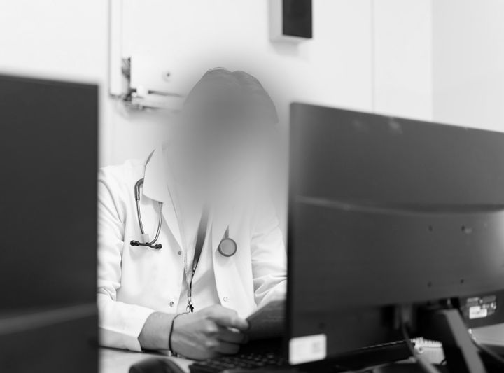 Une personne en blouse blanche, stéthoscope à la main, assise à un bureau avec deux écrans d'ordinateur, semble concentrée sur ses tâches administratives. Image en noir et blanc.