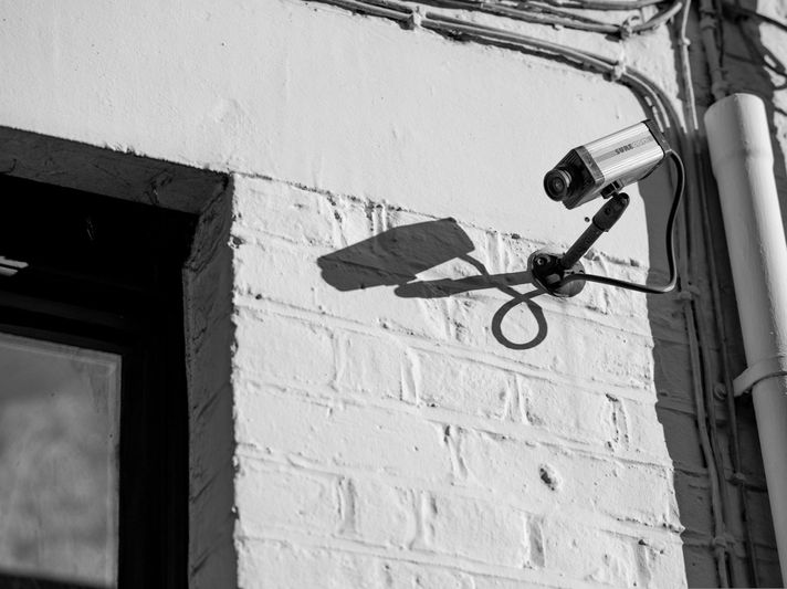 Imagen en blanco y negro de una cámara de seguridad instalada en una pared de ladrillos, proyectando una sombra. Cerca, se ven una ventana y algunos cables.