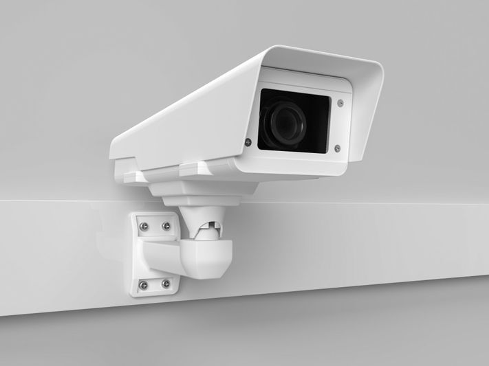 White security camera mounted on a gray wall, angled slightly downward, with a modern, rectangular design.