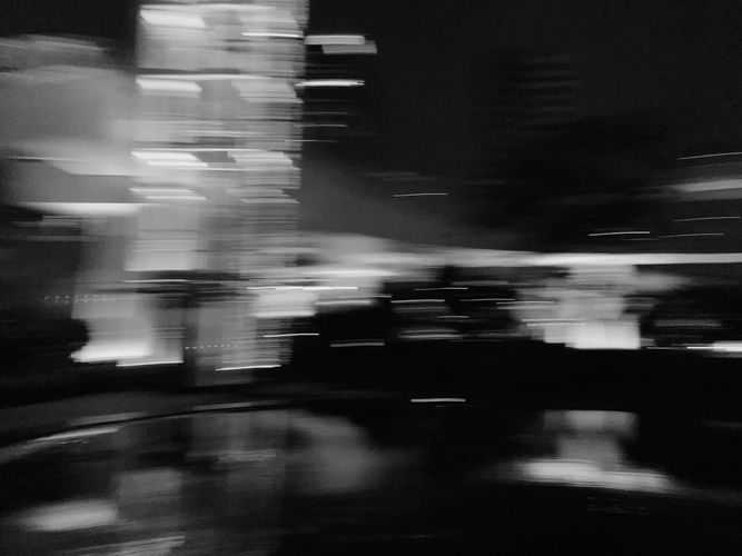 Blurry black and white image of city lights at night, with abstract light trails and reflections on a wet surface.