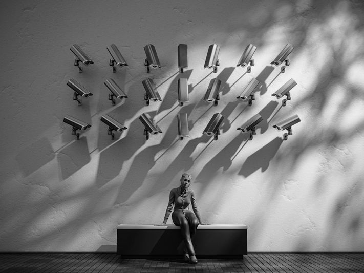 Person sitting on a bench under numerous surveillance cameras on a wall, with dramatic shadows cast in a black and white setting.