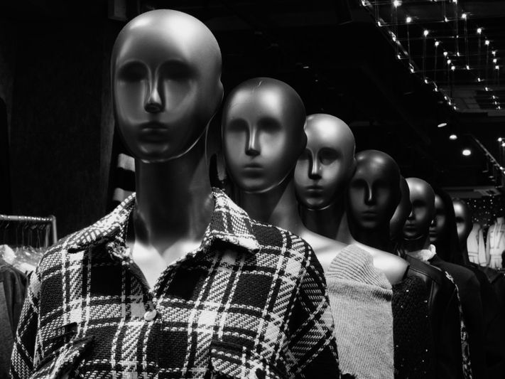 Rangée de mannequins dans un magasin, vêtus de tenues variées, avec un focus sur celui de devant portant une veste à carreaux. Image en noir et blanc.