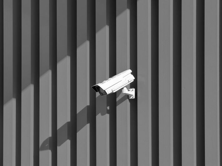 Security camera mounted on a corrugated metal wall, casting a shadow under angled sunlight.