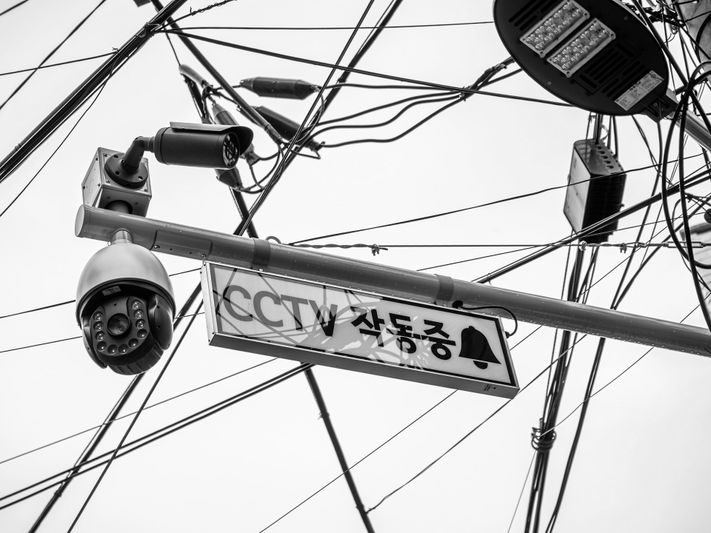 Image en noir et blanc de caméras CCTV montées sur un poteau avec un panneau coréen en dessous, entourées de nombreux fils aériens emmêlés.