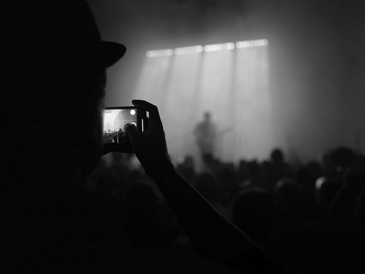 Silhouette of a person taking a photo with a smartphone at a concert, with a blurred musician on stage under bright lights.