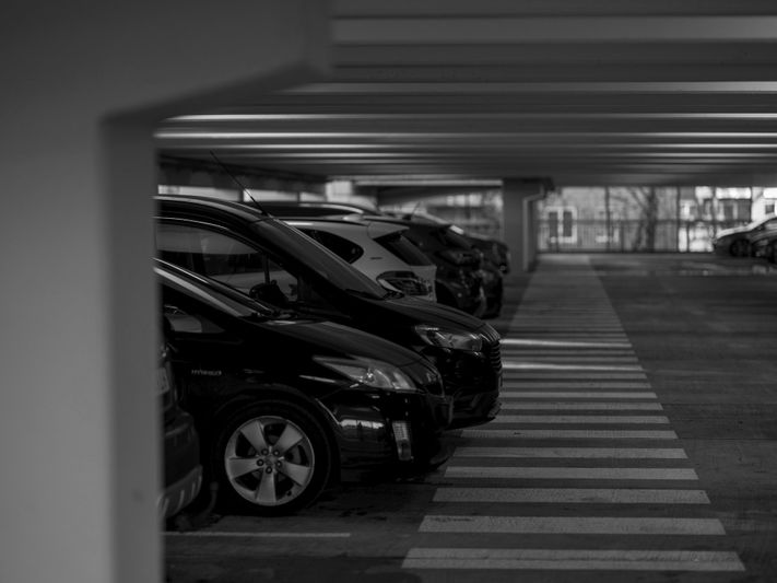 Image en noir et blanc de voitures stationnées dans un parking à plusieurs niveaux, avec des bandes au sol sur le béton et une lumière douce filtrant vers le bas.