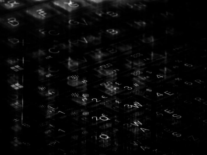 Close-up of a dimly lit keyboard with reflective keys displaying letters, numbers, and symbols in a dark, abstract setting.