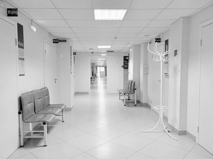 Una foto en blanco y negro de un pasillo de hospital con sillas vacías, paredes blancas y puertas a ambos lados, que conduce a un área abierta en la distancia.
