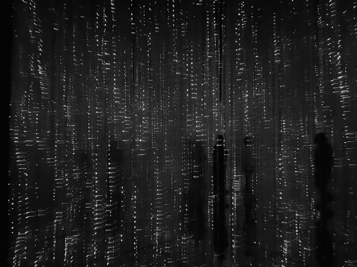 Image abstraite en noir et blanc de traînées lumineuses verticales ressemblant à de la pluie, avec des ombres de personnes en arrière-plan.