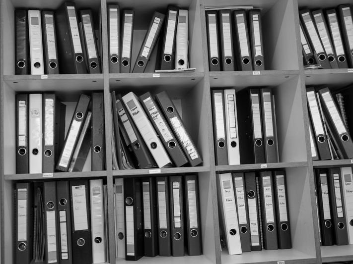 Rows of neatly organized ring binders on shelves, some leaning, in a monochrome setting.