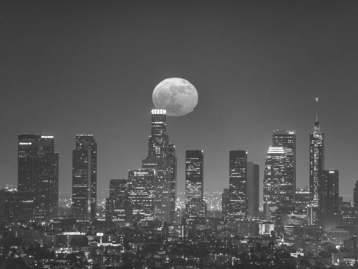 Photo en noir et blanc d'une silhouette de ville la nuit, avec une pleine lune placée directement au-dessus du plus haut gratte-ciel.