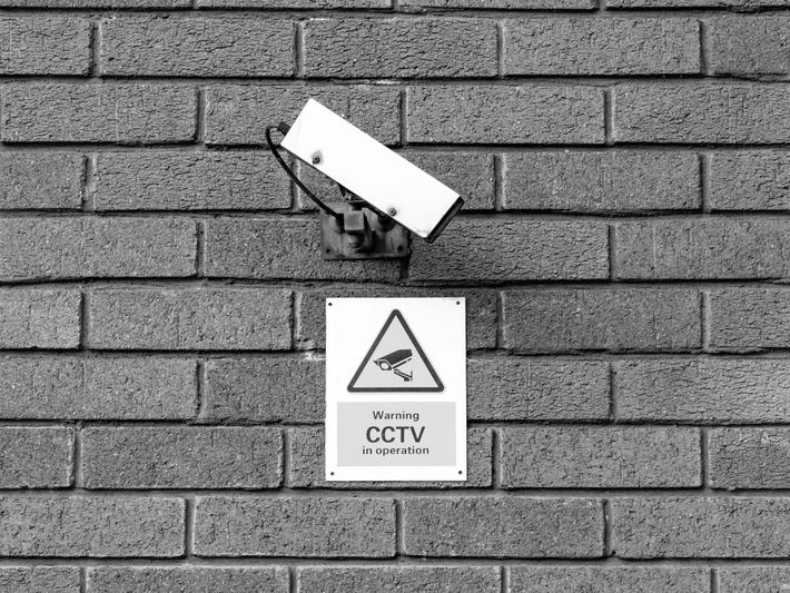 una cámara de vigilancia urbana blanca colgada en una pared de ladrillos, debajo de ella un cartel informativo que dice «Warning CCTV in operation»