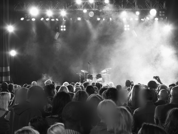 Black and white image of a concert stage with drums, bright lights, and a large crowd in the foreground.