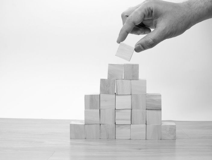 A hand places a wooden block on top of a pyramid-shaped stack of blocks on a wooden surface, against a plain background.
