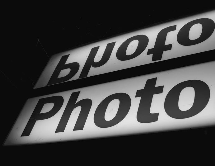 Primer plano en blanco y negro de un letrero luminoso con la palabra «Foto» y un reflejo invertido en un espejo encima.