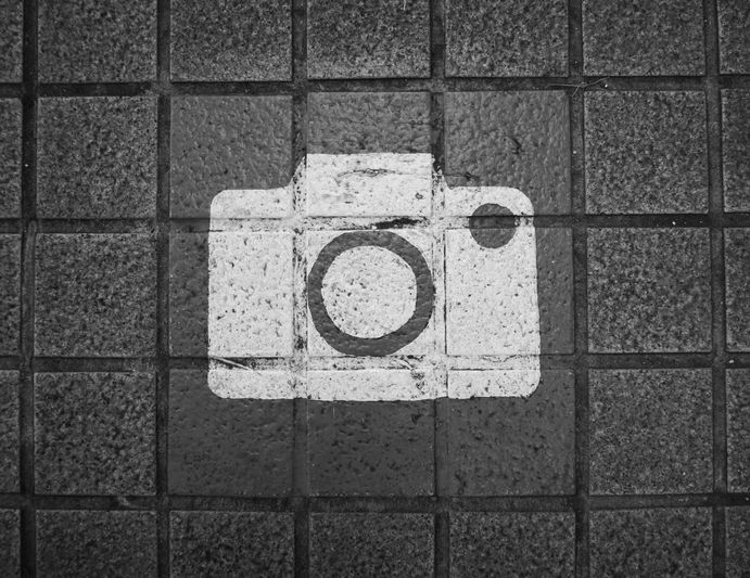 Ícono de cámara blanca pintado sobre mosaicos oscuros y texturizados, que simboliza un lugar o área para tomar fotografías.