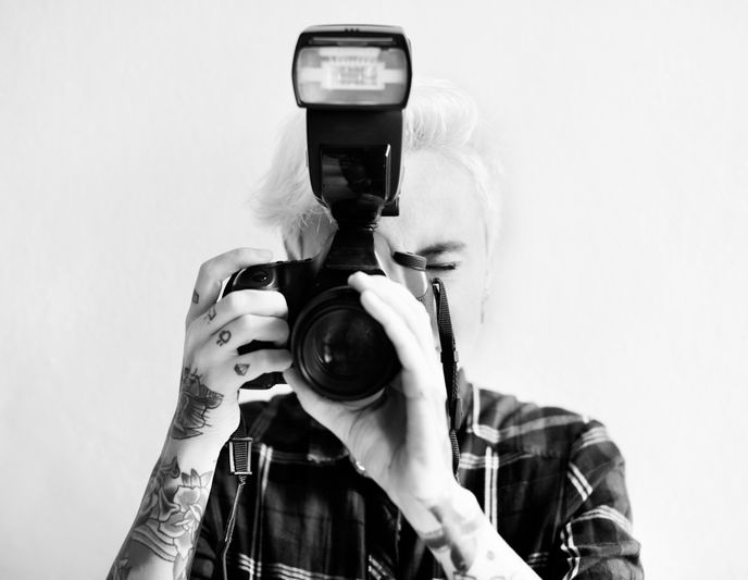 Personne tatouée tenant un appareil photo avec flash, vêtue d'une chemise à carreaux, sur un fond uni.