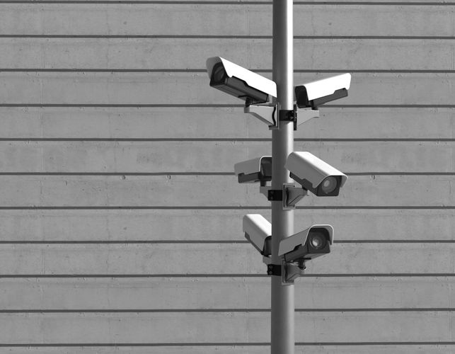 Five surveillance cameras mounted on a pole against a textured concrete wall background. Black and white image.