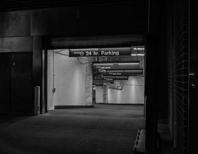 Photo en noir et blanc de l’entrée faiblement éclairée d’un parking ouvert 24 h/24, avec des panneaux de direction suspendus au plafond.